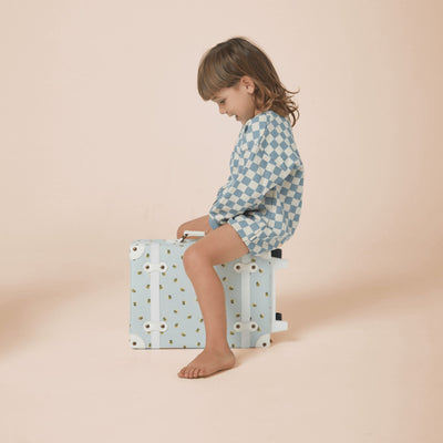 Child sitting on a pale blue and white bee print suitcase, wearing a blue checked sweater, from Olli Ella Kids’ See-Ya Suitcase collection