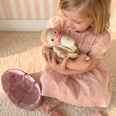 Young girl in a pink dress cuddles a Binky Dinky Moppet doll on a soft rug beside its pink carrycot from Dinkum Dolls by Olli Ella