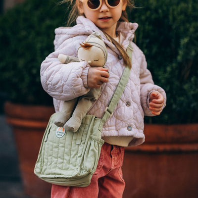 Child carrying a soft doll and wearing a sage green quilted changing bag by Dinkum Dolls, part of a playful on the go doll accessory set