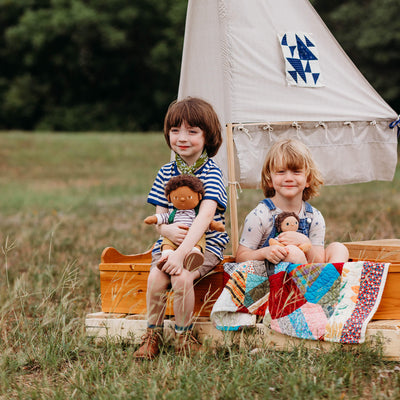 Two children sit outdoors beside a small tent, cuddling dolls beneath a colorful patchwork blanket in a grassy field.
