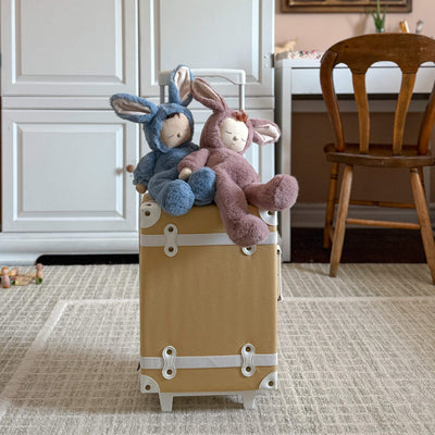 Two Cozy Dinkums bunny plush toys, one blue and one mauve, sit atop a tan vintage-style suitcase in a cozy home interior.