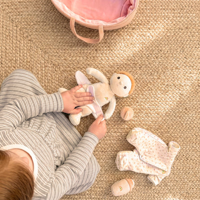 Child playing with a small cream-colored Binky Dinky doll on a woven rug, with striped pajamas, floral clothing, accessories, and a pink carry bag nearby.