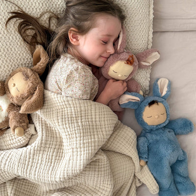 Child cuddling Cozy Dinkums Bunny Twiggy, a soft pink bunny doll, in bed with two other plush animal dolls under a textured cream blanket.