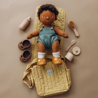 Dinkum Dolls doll in a teal romper and orange boots rests on a honey quilted changing mat with a bottle, shoes, brush, and care accessories nearby