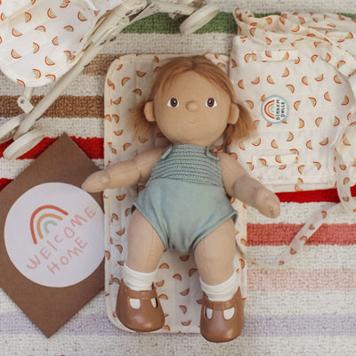 Dinkum Dolls toy doll lying on a rainbow print changing mat, wearing a blue romper and brown shoes, with matching accessories and a Welcome Home card nearby