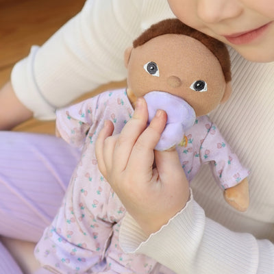 A child cuddles Binky Dinky Pip, a soft brown haired doll in a floral dress with a lavender pacifier from Dinkum Dolls by Olli Ella