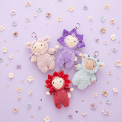 Plush Dinky bag charms in pink, purple, blue, and red, including Rose with a friendly face and flower outfit, displayed among pastel flowers on a lavender background