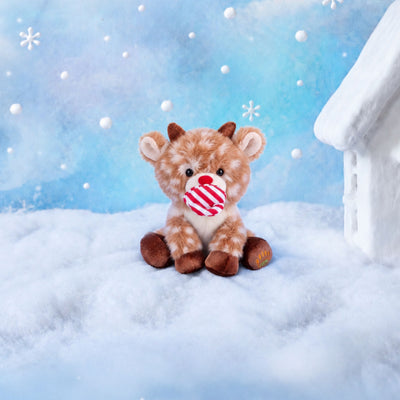 Fluffy plush reindeer with fawn fur and cream spots, a red nose, a smiling cream muzzle, short brown corduroy antlers and hooves, and an embroidered paw-and-rainbow motif on one foot, sitting beside a red and white striped hat.