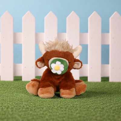 Dinkum Pets Binky Cow Gus plush Highland cow toy seated on green grass, with brown fur, cream horns and mane, and a green and white binky.