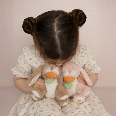 Child in a floral dress cuddles two soft bunny toys, one brown and one pink, each with a colorful pacifier, from Dinkum Pets Binky Bunny Dot.
