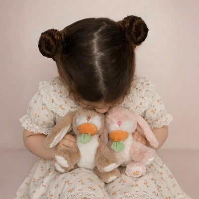 Child in a floral dress cuddles two soft plush bunnies, one tan and one pink, each holding a colorful carrot, from Dinkum Pets.