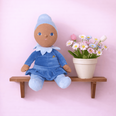 Dinky Dinkums Iris plush doll in a blue dress and matching cap sits on a wooden shelf beside a potted bouquet of colorful flowers against a pink background