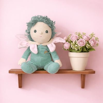 Dinky Dinkums Lily plush doll in a pale green romper with pink wings, seated beside a potted bouquet of pink flowers on a wooden wall shelf against a pastel pink background.