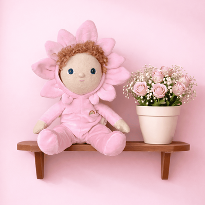 Dinky Dinkums Magnolia doll in a soft pink flower costume sits on a wooden wall shelf beside a potted bouquet of pink roses against a pastel pink background by Dinky Dinkum Dolls