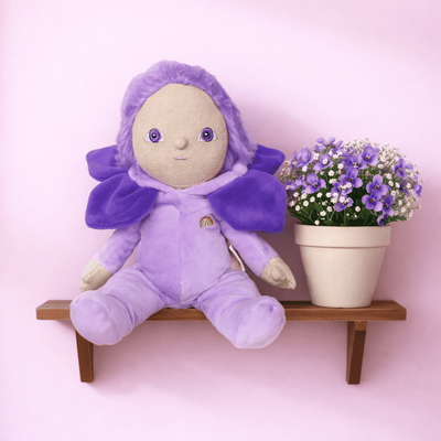 Purple Dinky Dinkum plush doll with violet hair and a petal cape sits on a wooden shelf beside a potted bouquet of purple and white flowers.