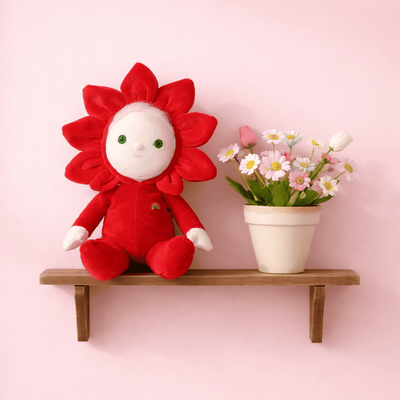 Red Dinky Dinkum Dolls flower doll with a smiling face sits beside a potted bouquet of pink and white flowers on a wooden wall shelf.