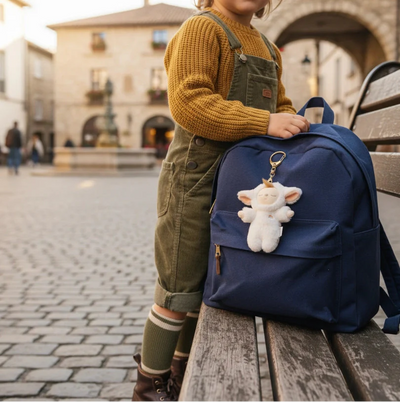 White lamb plush bag charm clipped to a navy backpack resting on a park bench, with a child in overalls standing beside it in a cobblestone square