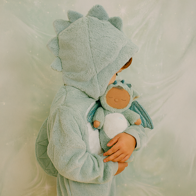 A child in a pale green dragon costume cuddles a Cozy Dinkum Dragon Ash plush toy with teal wings and a warm orange face.