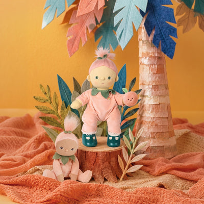A Dinkum doll wears a peach romper with a green collar and teal shoes while holding a peach toy beside a smaller doll in a colorful jungle scene by Dinkum Dolls by Olli Ella