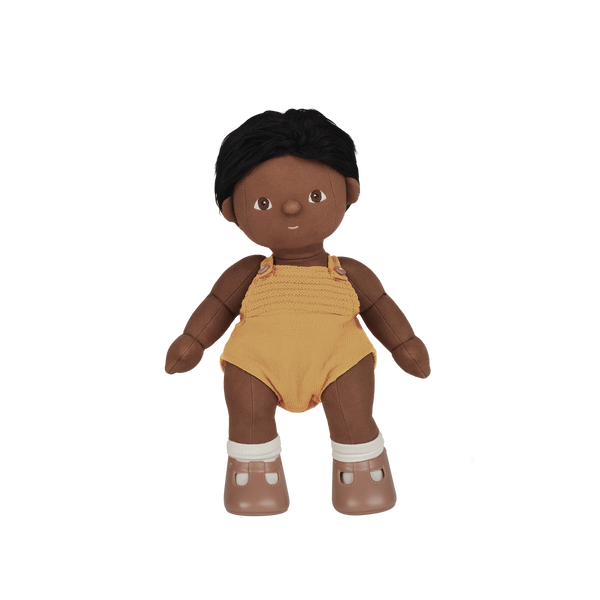 Dinkum Dolls Tiny doll with dark skin, short black hair, a mustard yellow romper, white socks, and brown Mary Jane shoes standing against a black background