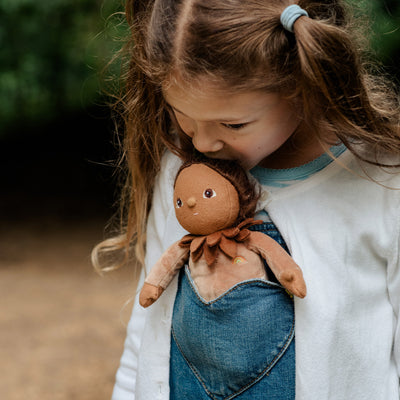 A child cuddles a Dinky Dinkums Forest Friends Allie Acorn soft doll tucked into a denim pocket, showcasing its brown acorn inspired design and leaf collar.