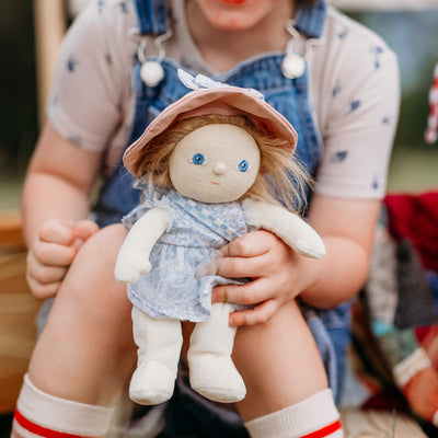 Child holding a soft Dinky Dinkum doll with blue eyes, blonde hair, a blue floral dress, and a pink sunhat.