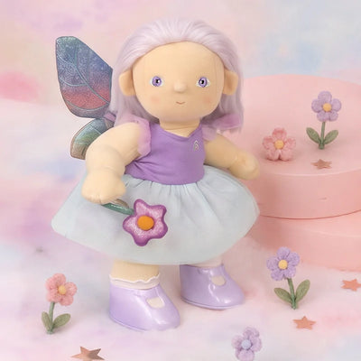 Fairy Evie Dinkum doll with lavender hair, purple top, white skirt, glittery wings and matching boots surrounded by colorful flowers on a pastel background