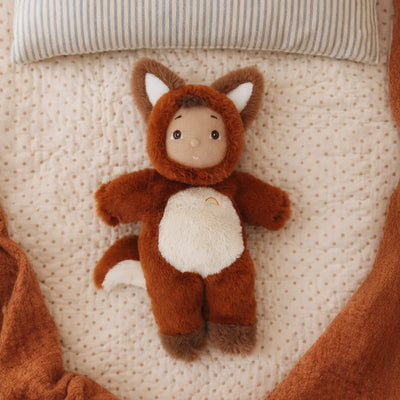 Cozy Dinkums Freddy Fox plush doll in a rust colored fox costume lying on a cream quilted bed beneath a striped pillow