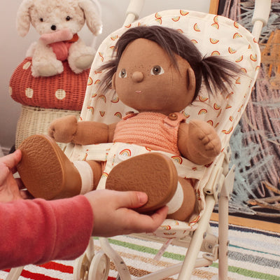 Dinkum Dolls doll seated in a rainbow print stroller seat, wearing a peach knit outfit while a child adjusts the doll’s shoes indoors