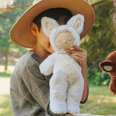 A child in a straw hat cuddles a soft white fox plush toy outdoors, showing its sleepy embroidered face and fluffy ears.
