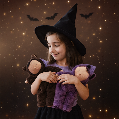 Child wearing a black witch hat cuddles two bat plush dolls, one brown and one purple, against a sparkling night sky with silhouetted bats.