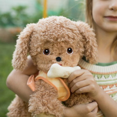 Child cuddling Goldie, a soft golden Dinkum Dogs plush puppy holding a white bone treat in its mouth outdoors