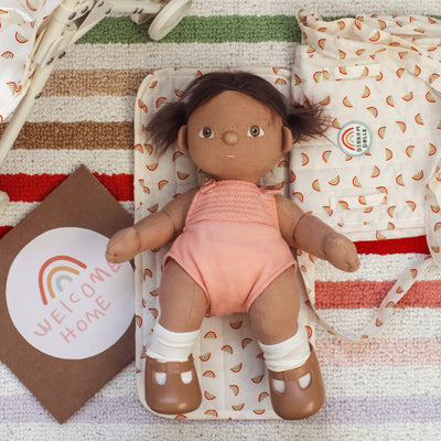 Dinkum Dolls doll with brown skin, soft brown hair, pink romper, white socks and brown shoes lying on a rainbow print blanket beside a welcome home card