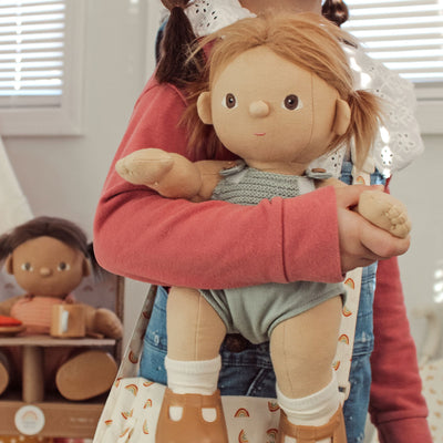 Child holding a soft Dinkum Dolls doll with golden hair, a powder blue romper, white socks and brown boots, while another doll sits in the background during pretend play