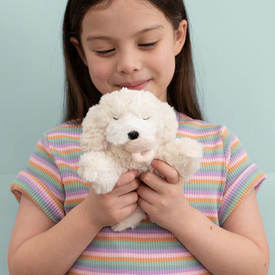 Child cuddling a soft cream Binky Puppy plush toy with shaggy fur, floppy ears, and a black nose from Dinkum Pets