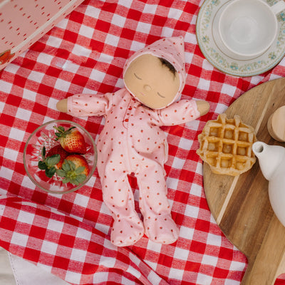 Dozy Dinkums Pip Strawberry doll in a pink strawberry print outfit lying on a red gingham picnic cloth beside fresh strawberries and a waffle