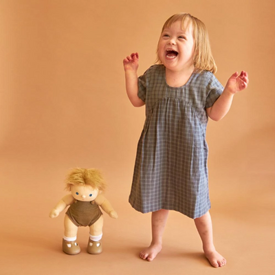 Smiling child standing beside a blonde Dinkum Dolls Poppet doll in a brown outfit, both pictured against a warm peach background for playful dress up fun