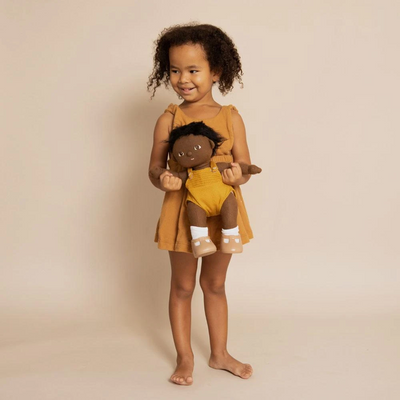 Child holding a dark skinned Dinkum Dolls Tiny doll in a mustard romper and white shoes, smiling while standing barefoot in a warm studio setting