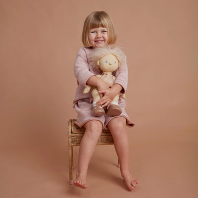 Smiling child in a pink dress cuddles a Dinkum Dolls Petal doll while seated on a wicker chair, capturing imaginative play and companionship.