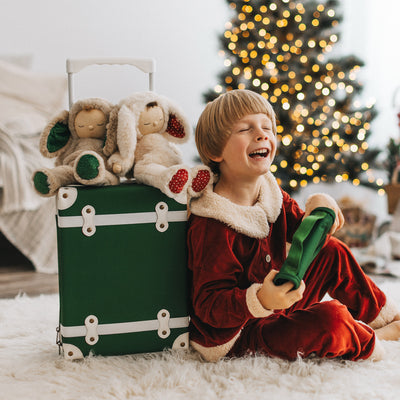Child in red velvet pajamas holds a green backpack beside a Forest See-Ya Suitcase filled with plush toys, photographed by a glowing Christmas tree for Olli Ella Kids