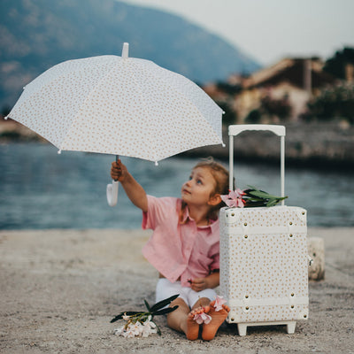 Olli Ella Kids children’s umbrella with a cream dotted canopy held by a child beside a matching suitcase on a lakeside beach rescue outfit