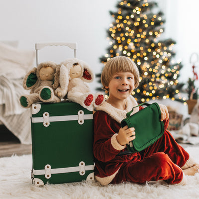 Child in festive red pajamas holds a green See-Ya wash bag beside a matching suitcase topped with plush toys and a glowing Christmas tree from Olli Ella Kids