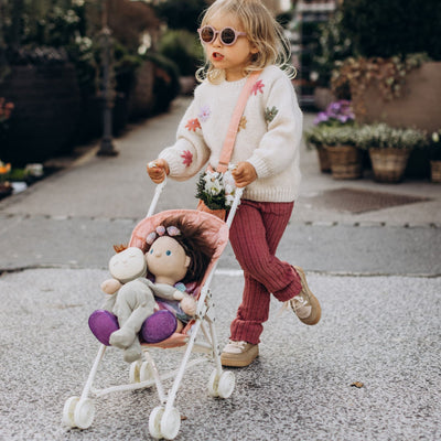 Child wearing sunglasses pushes a white doll stroller with a rose pink seat and two dolls inside outdoors, featuring a Dinkum Dolls Sollie stroller seat