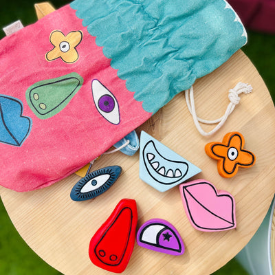 Colorful facial expression sensory stones with eye, mouth, nose, and lip shapes beside a pink and turquoise drawstring pouch on a wooden surface