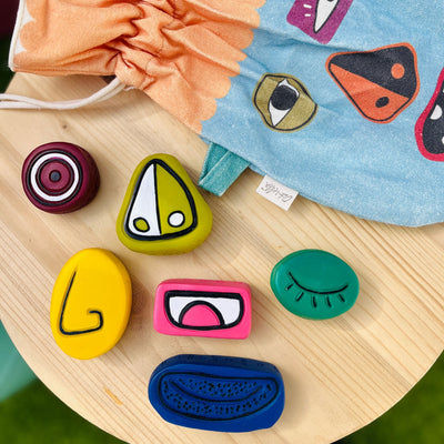 Colorful Silly Sam sensory stones with interchangeable facial features arranged on a wooden table beside a patterned blue play bag and orange drawstring pouch by Olli Ella Kids