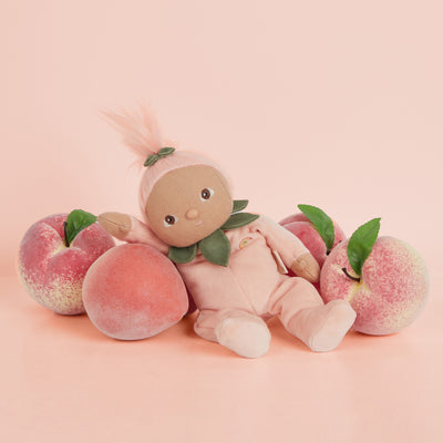 Dinky Dinkums Peggy Peach doll in a peach-colored outfit and leafy collar, reclining among fresh peaches against a soft pink background