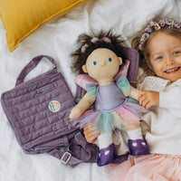 Dinkum Dolls lavender changing set with a soft doll in a colorful dress, quilted purple carry bag, and smiling child relaxing together on a bed