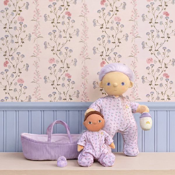 Two dolls in matching outfits with a baby basket against a floral wallpapered wall.