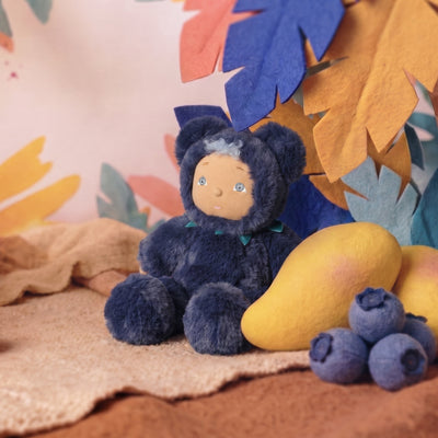 A plush doll dressed in a dark navy blue teddy bear outfit sits on a beige blanket, styled with a yellow mango and blueberries against a colorful felt leaf backdrop.