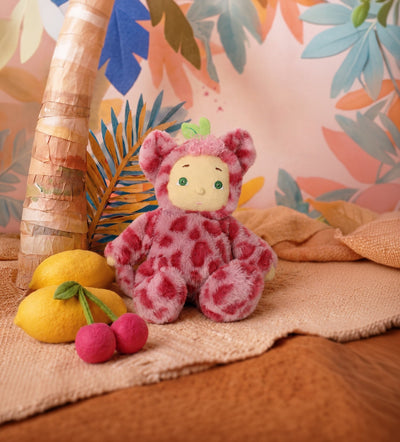 A plush teddy bear doll wearing a pink spotted onesie and a small green leaf on its head, posed on a beige blanket with lemons, cherries, and a palm tree against a tropical leaf patterned backdrop.